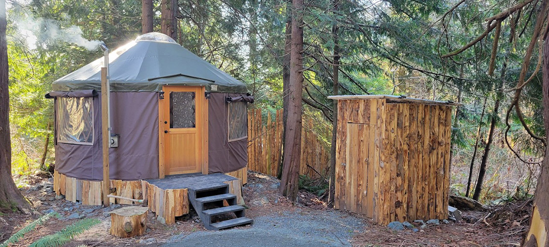 Photo of the Sea to Tree yurt from the outside; A round structure surrounded by trees, and an outhouse next to it.