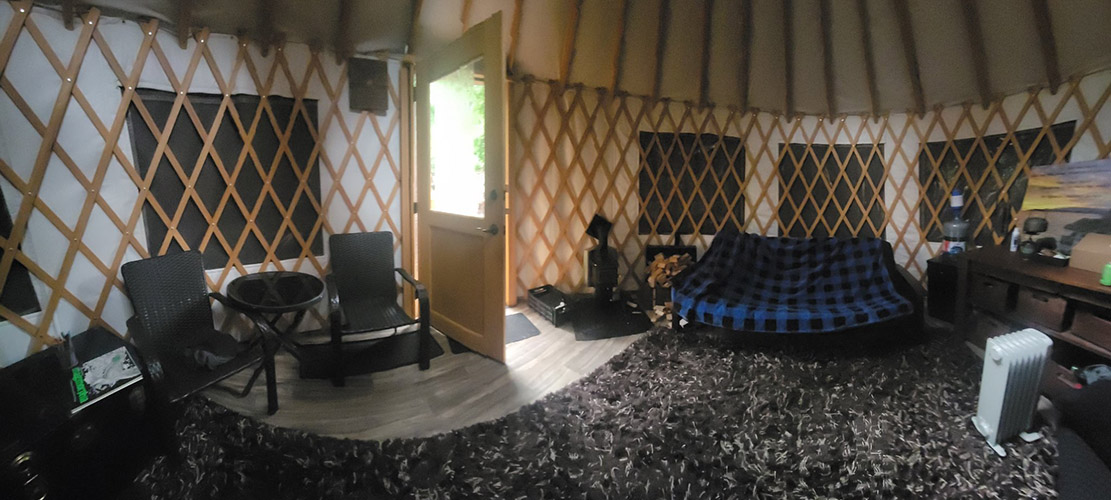 Photo of the inside of the Sea to Tree yurt; You can see armchairs, a sofa, coffee table, a wood stove and a space heater.